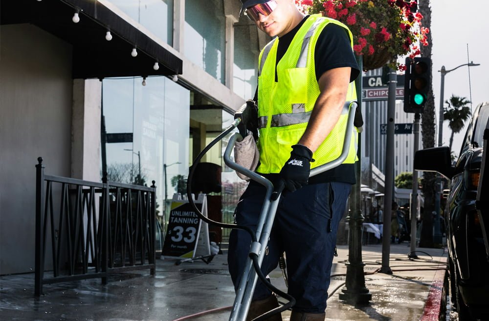 Worker in safety gear using a pressure washer for commercial exterior cleaning on a sidewalk, showcasing professionalism and commitment to cleanliness.