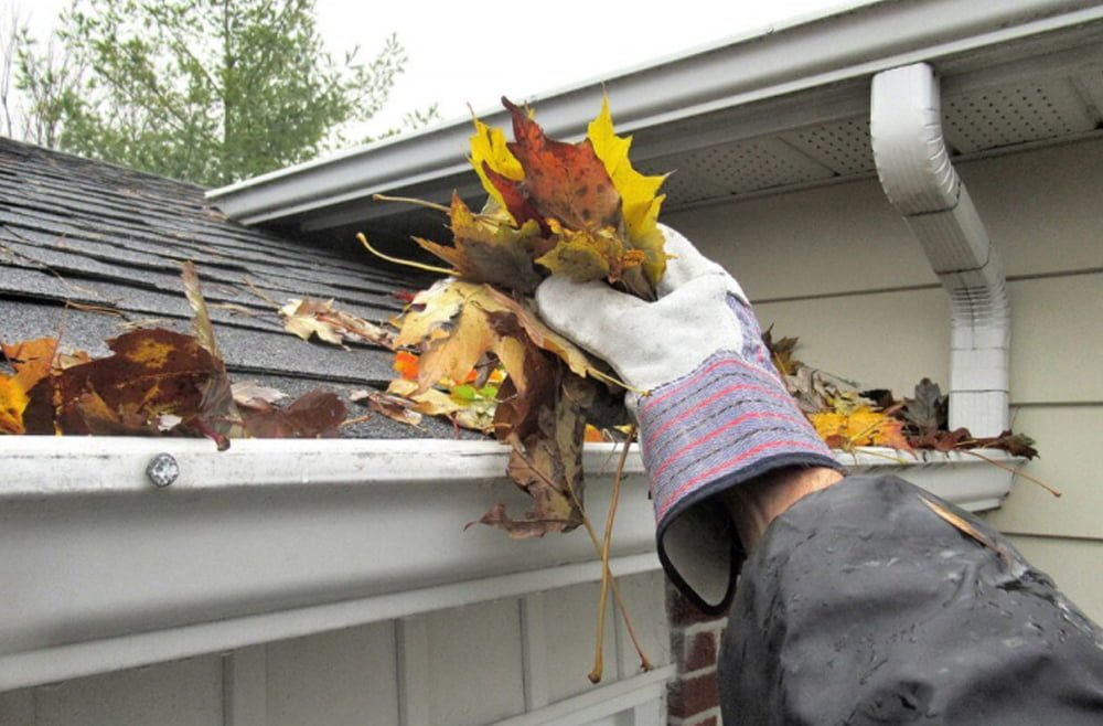 Person wearing gloves cleaning leaves from a residential gutter, highlighting the importance of gutter maintenance for preventing water damage, relevant to Frater Exterior Cleaning Services.