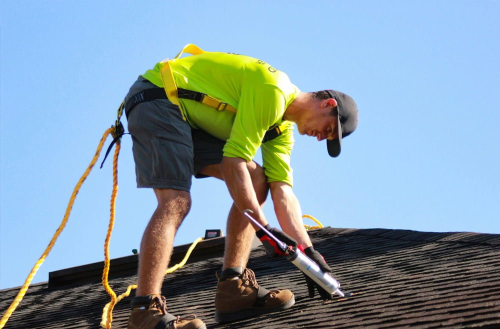Worker applying roof treatment to remove moss and algae, wearing safety harness, bright yellow shirt, and shorts, against clear blue sky.