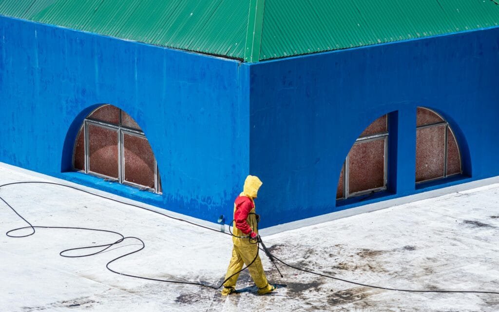 A worker in protective gear power washes a vibrant blue building exterior.